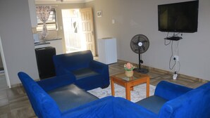 Un salon avec un canapé bleu, une petite table basse en bois sur laquelle est posé un vase, un ventilateur sur pied et un téléviseur fixé au mur.
