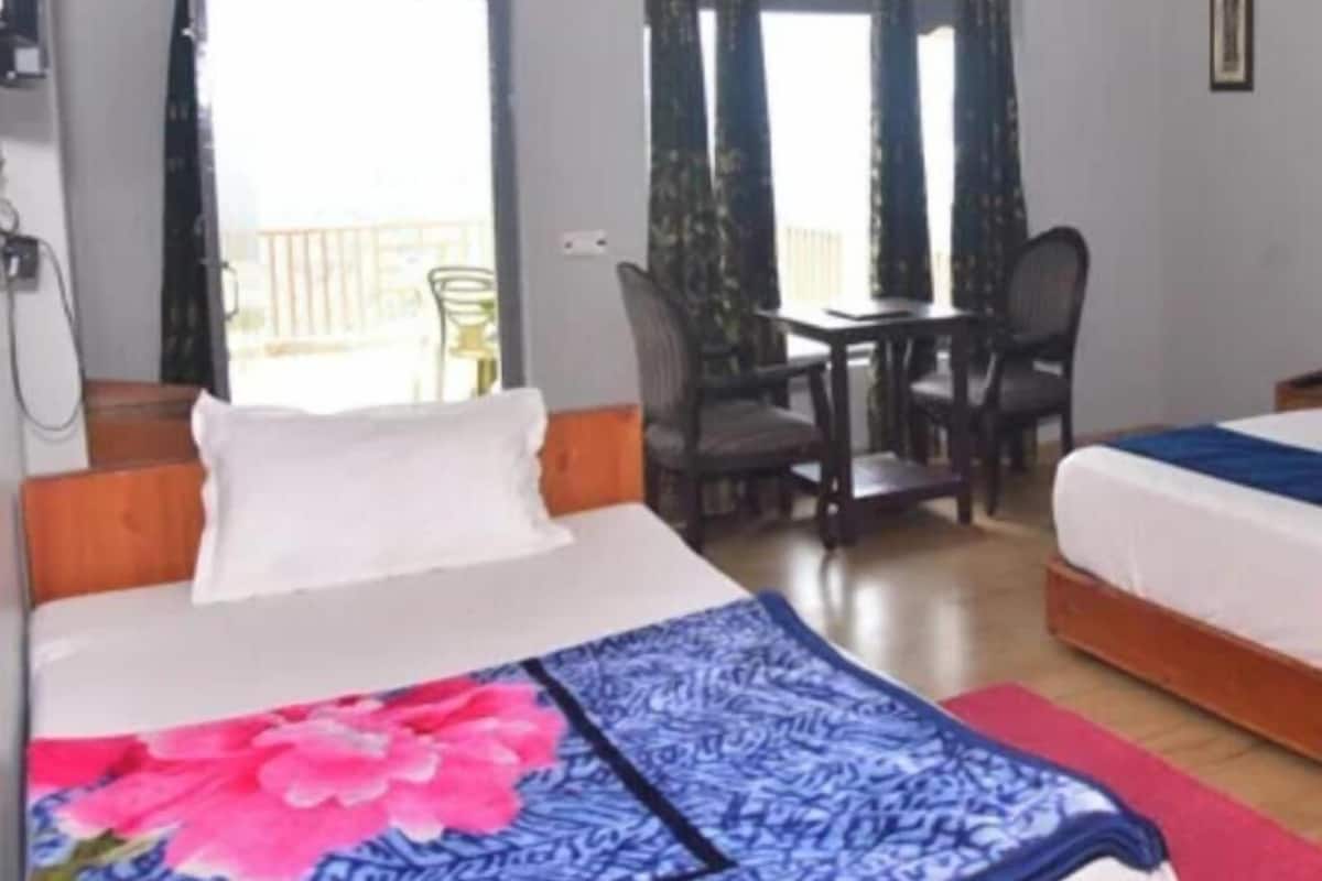 Quarto de hotel com cama, escrivaninha e cadeira. Há uma janela com cortinas, e é visível uma varanda lá fora.