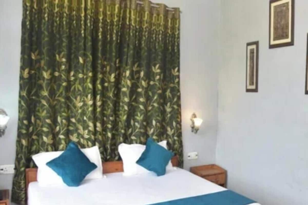 Quarto com cama grande, cabeceira de madeira, colcha azul e cortina verde com folhagem.