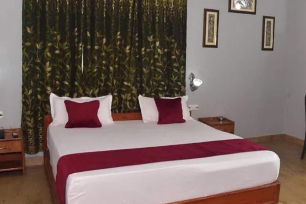 Uma cama de casal com colcha branca e vermelha, duas almofadas brancas e um cobertor vermelho. Há uma mesa de cabeceira de madeira à esquerda, um abajur à direita e uma planta ao fundo.