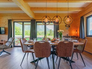 Un intérieur en bois avec une table à manger dressée pour quatre personnes, entourée de chaises, et une grande fenêtre offrant une vue sur la verdure et les montagnes.