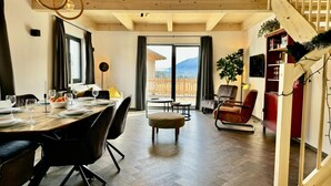 A spacious room with a wooden ceiling, a dining area with a table set for four, a comfortable seating area with a sofa and armchairs, and a view of a wooden fence and mountains through large windows. - Beautiful chalet near the Nassfeld skiing area (Obermöschach)