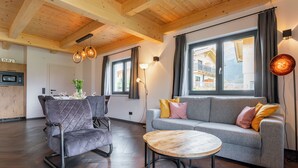 A modern living room with a grey sofa, a round wooden coffee table, and a grey armchair. There is a dining area with a white table and chairs, and a kitchen with a microwave. Large windows provide natural light and a view of the outside. - Beautiful chalet near the Nassfeld skiing area (Obermöschach)