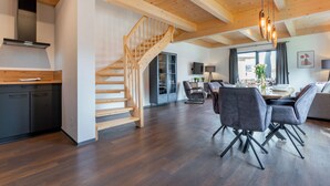 A spacious living area with a wooden staircase, a dining table with chairs, and a kitchen area with a modern stove.