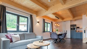 A modern living room with a grey sofa, round wooden coffee table, and a dining area with a wooden table and chairs. A kitchen with dark cabinets and a wooden ceiling is visible in the background.