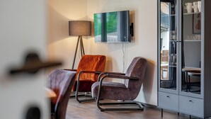 Een moderne woonkamer met een bank, een fauteuil, een vloerlamp, een televisie en een kast met glazen deuren.