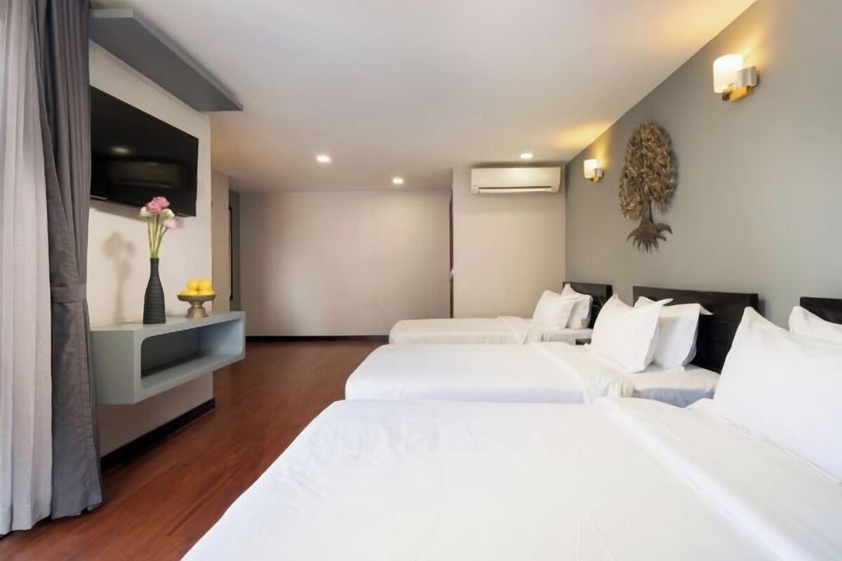 Une chambre d’hôtel avec trois lits, un téléviseur à écran plat, un climatiseur fixé au mur et un arbre décoratif sur le mur.