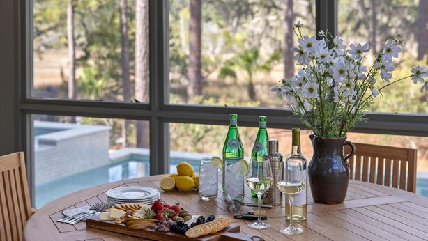 A dining table with a variety of food and drinks, a vase of flowers, and a view of trees through large windows. - Luxe Interiors + Pool and Views overlooking Marsh (Bluffton)