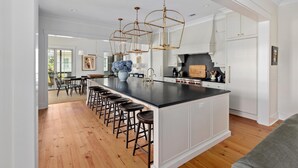 A modern kitchen with a large island, bar stools, pendant lighting, and white cabinetry. - Luxe Interiors + Pool and Views overlooking Marsh (Bluffton)