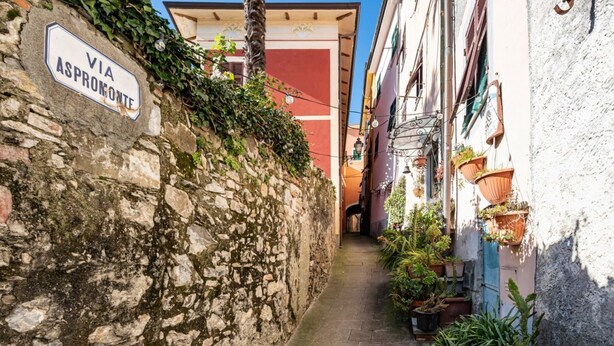 A narrow alleyway with a stone wall, a sign reading 'VIA ASPROMONTE,' and potted plants along the path.