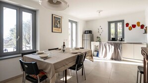 A dining area with a table set for four, a kitchen in the background, and large windows.