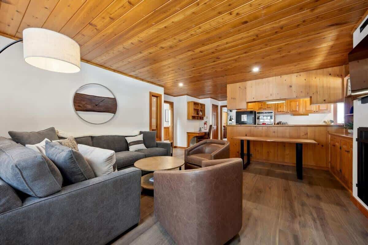 A modern interior with a wooden ceiling, a living area with a sofa and armchairs, a kitchen with wooden cabinets, and a round wall clock.