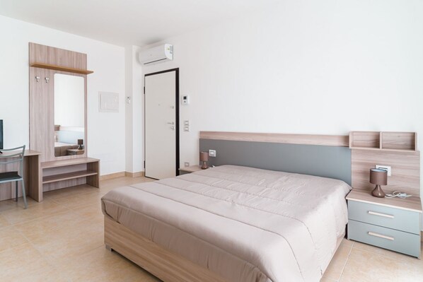 A bedroom with a bed, a desk, a chair, a wardrobe, a mirror, a nightstand, and a wall-mounted air conditioner.