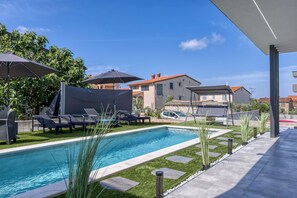 Ein rechteckiger Pool mit Fliesenrand, umgeben von einer Terrasse mit Liegestühlen und Sonnenschirmen.