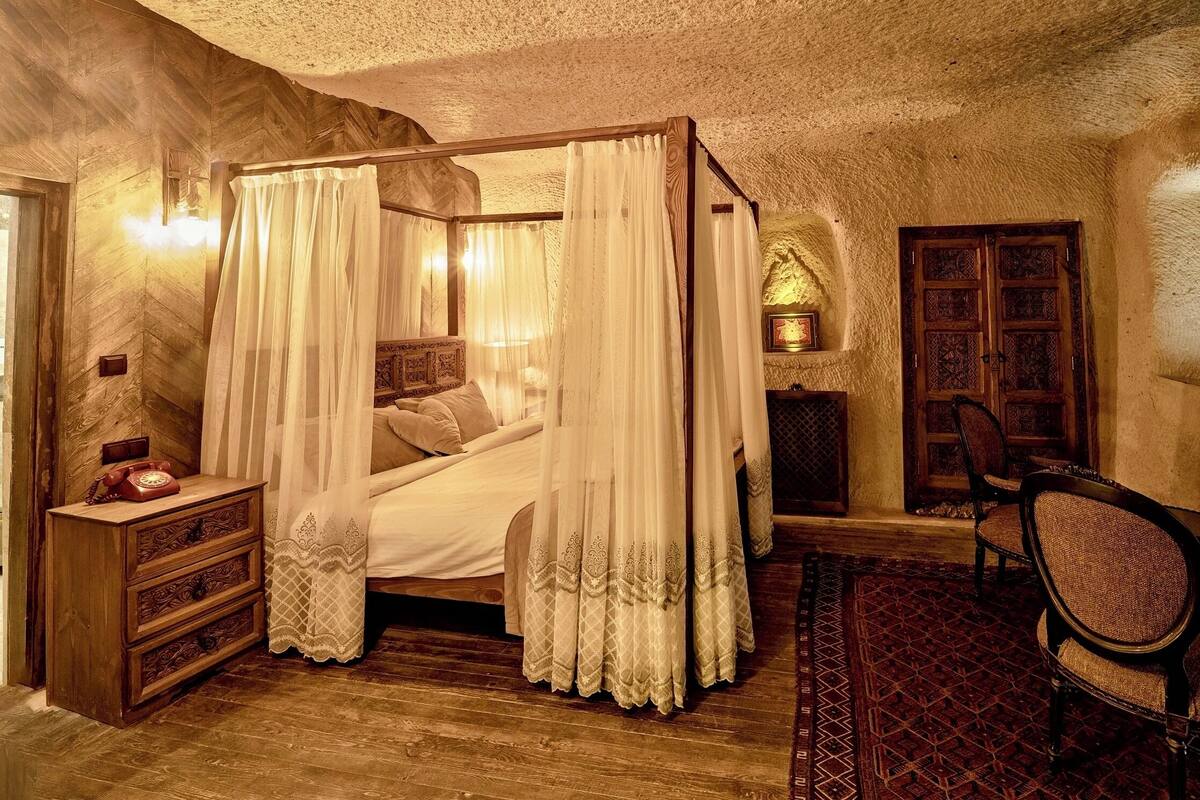 A four-poster bed with white linens and a canopy, a wooden nightstand with a rotary phone, a patterned rug, and a traditional wooden door.
