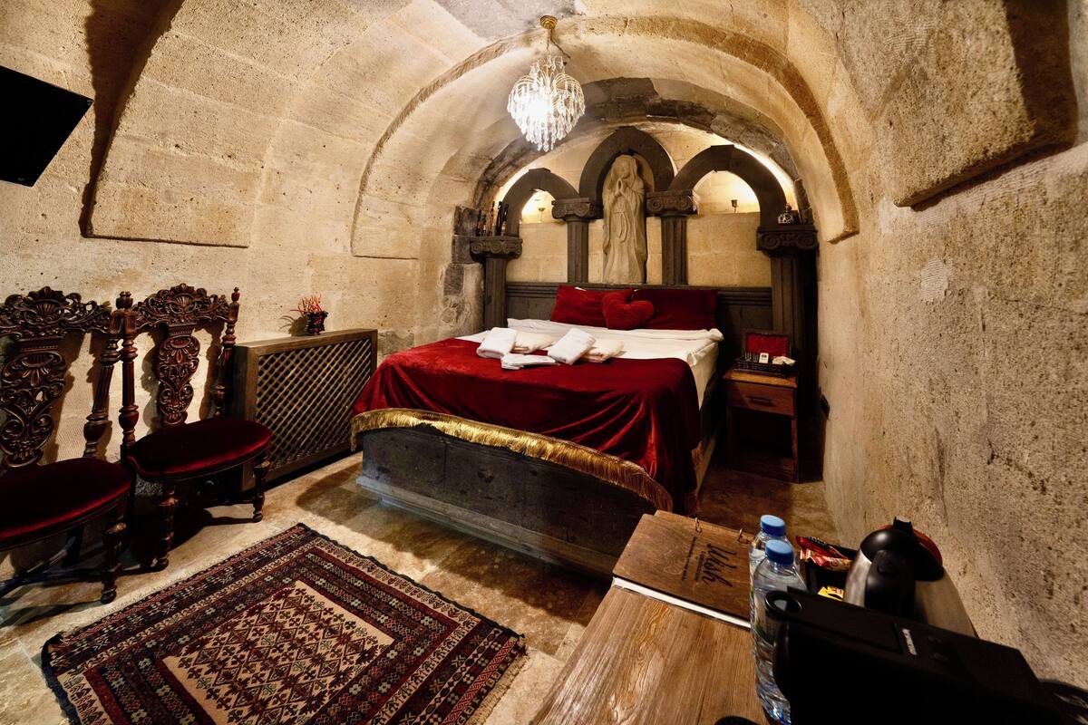 A bedroom with a stone arched ceiling, a bed with red bedding, a wooden chair, a small table, and a chandelier.
