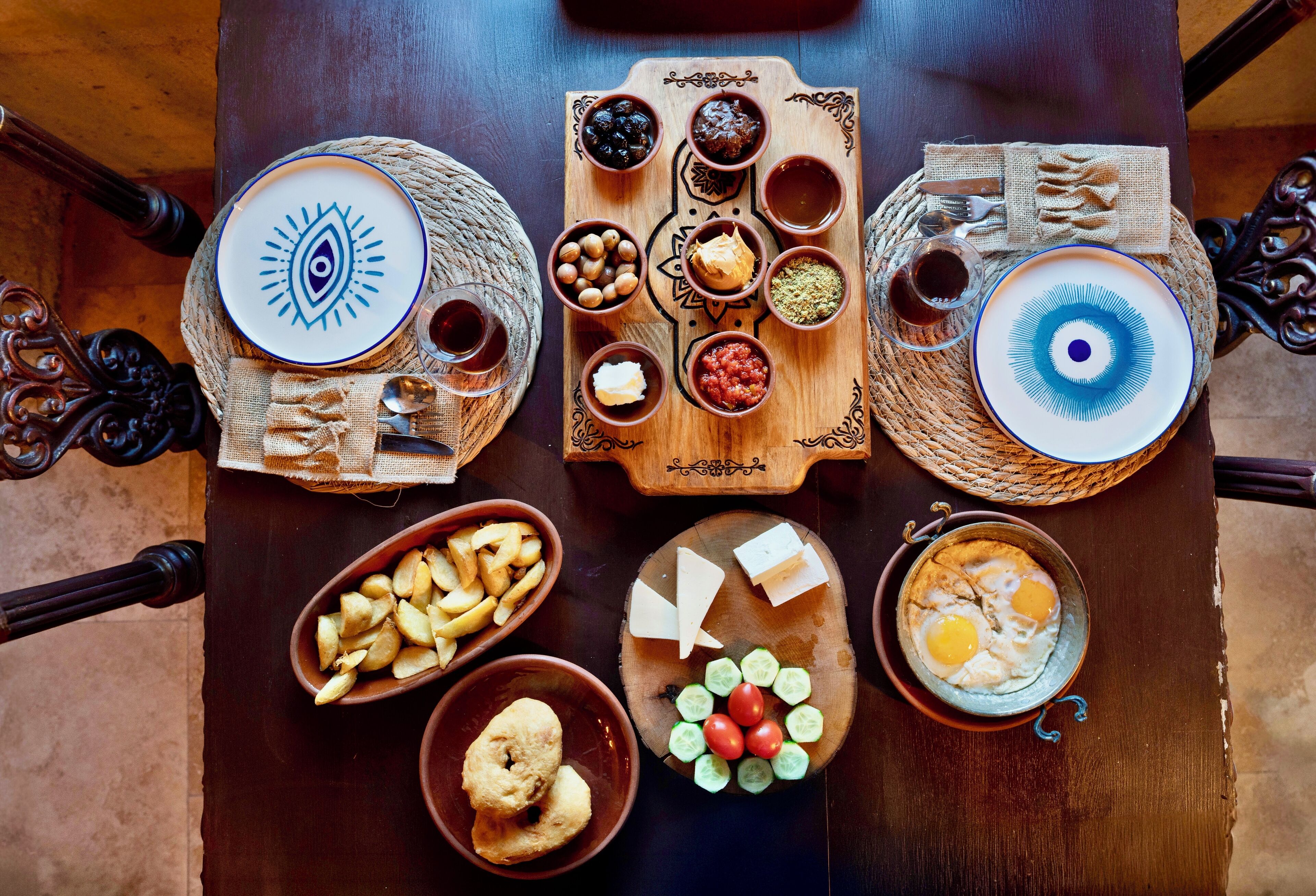 A wooden table with various food items and dishes, including bowls of nuts, cheese, and sliced cucumbers, as well as plates with eggs and bread.
