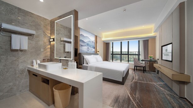 A modern hotel room with a large bed, a separate desk, a bathroom with a sink and mirror, and a view of the outdoors. - Guangzhou Nansha Meihao Lizhi Hotel (Guangzhou)