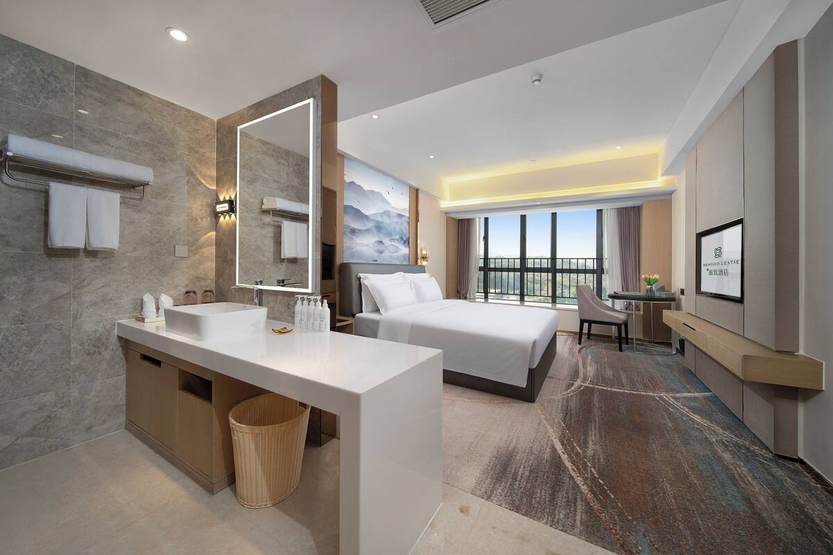 A modern hotel room with a large bed, a separate desk, a bathroom with a sink and mirror, and a view of the outdoors.
