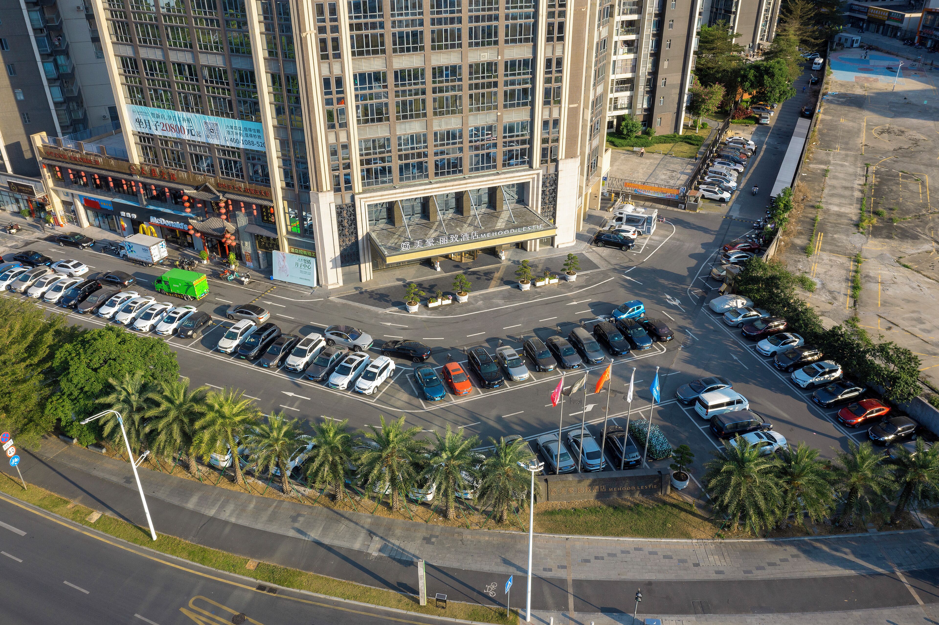 A multi-story hotel with a modern glass facade, a parking lot with various vehicles, and a street with palm trees.