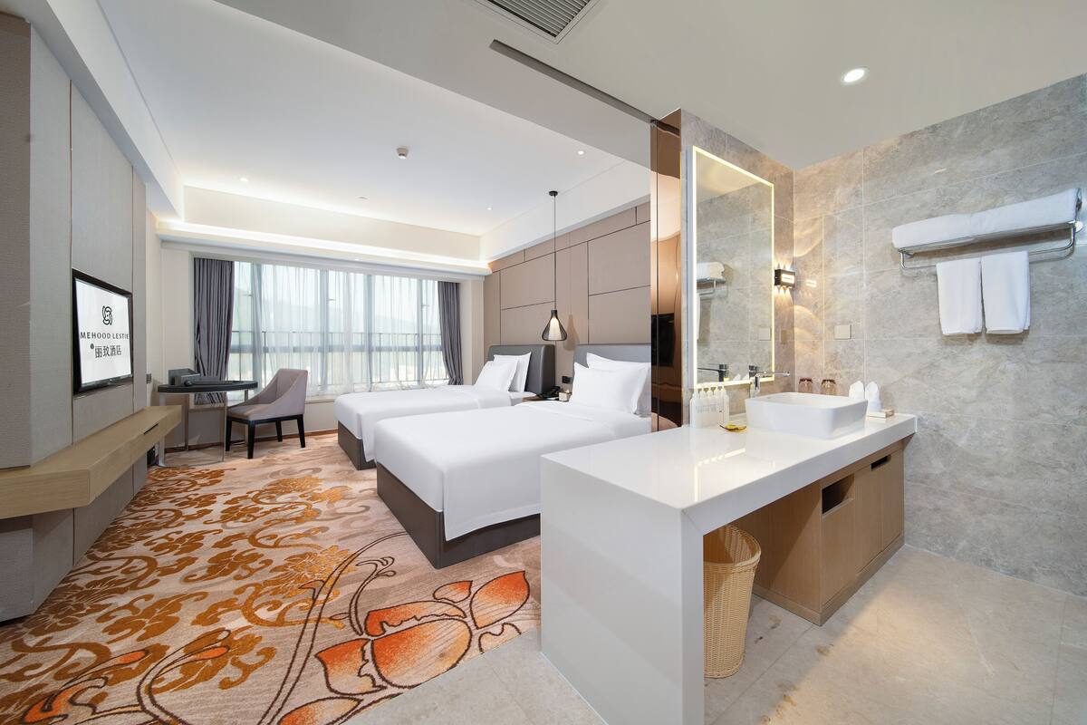 A hotel room with a large bed, a desk, a chair, a bathroom with a sink and mirror, and a large window with curtains.