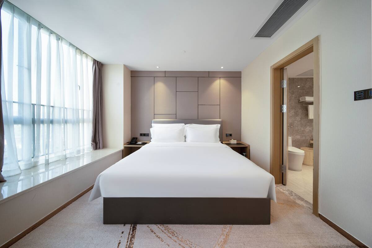 A modern hotel room with a large bed, bedside tables, a window with curtains, and a view of the bathroom.