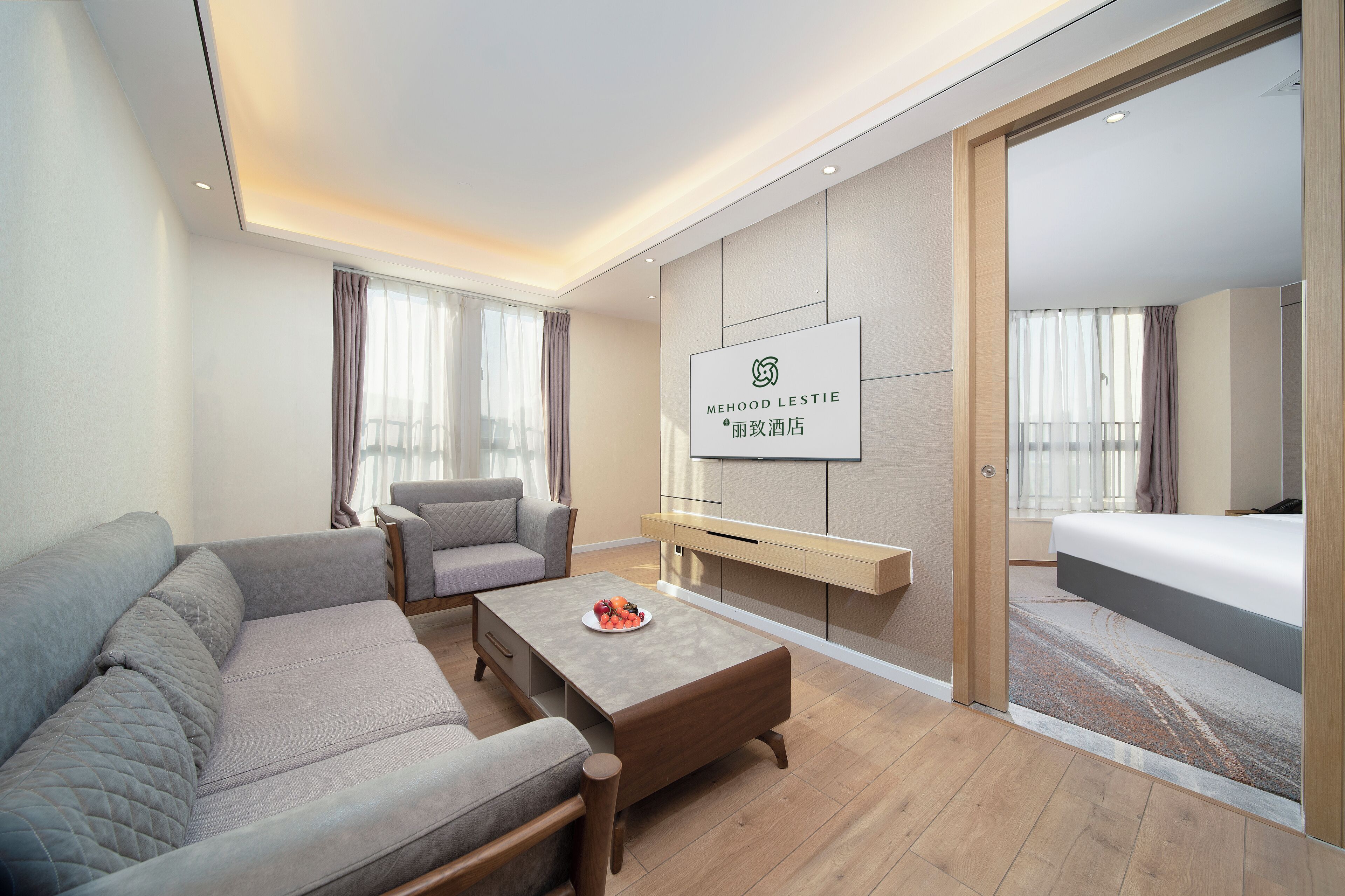 A modern hotel room with a sofa, a coffee table with fruit, a TV, and a bed.