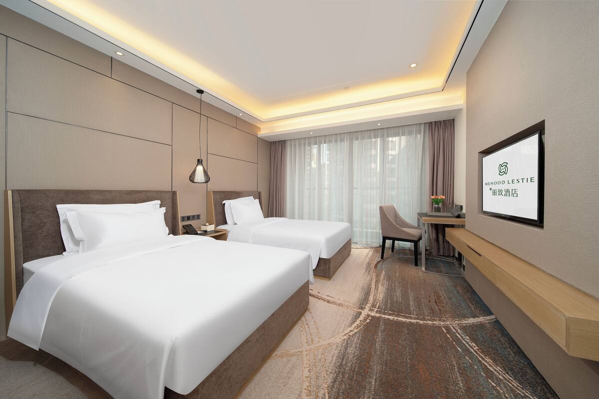 A hotel room with two beds, a desk, a chair, a TV, and a large window with curtains.