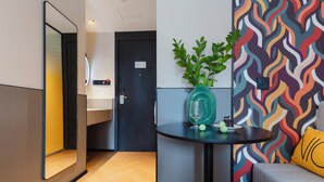 Une chambre d’hôtel moderne avec un plancher en bois, une porte noire, un miroir, une petite table avec un vase et un verre, et un mur aux motifs ondulés et aux couleurs vives.