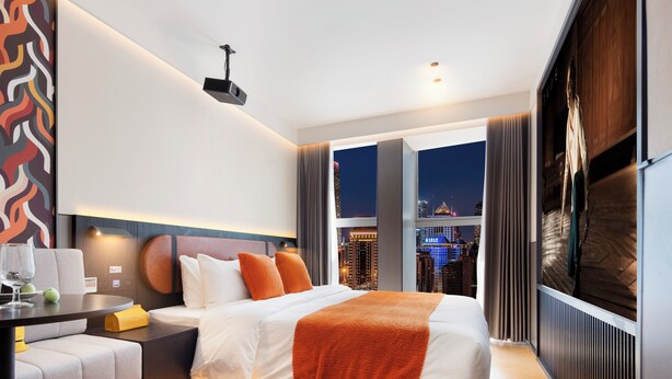 Une chambre d’hôtel moderne avec un grand lit, un bureau et une vue sur le paysage urbain illuminé la nuit.