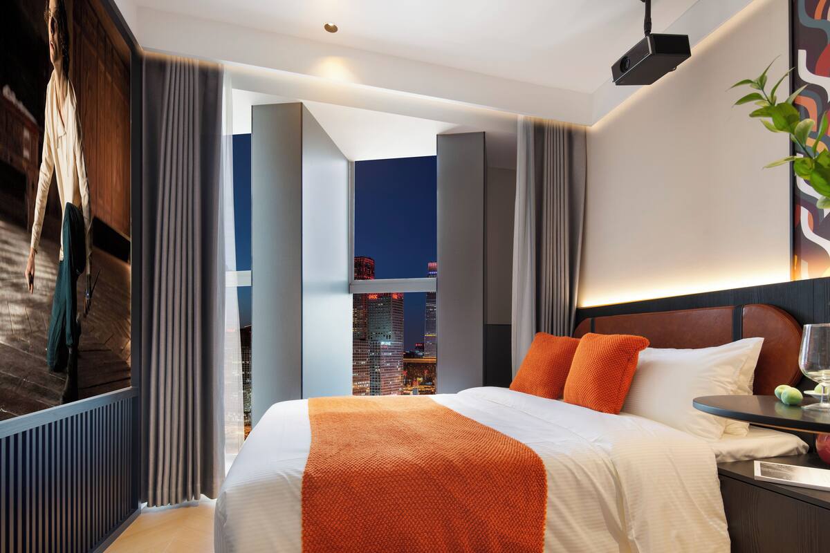 A modern hotel room with a large bed, a view of the cityscape, and a city skyline visible through the window.