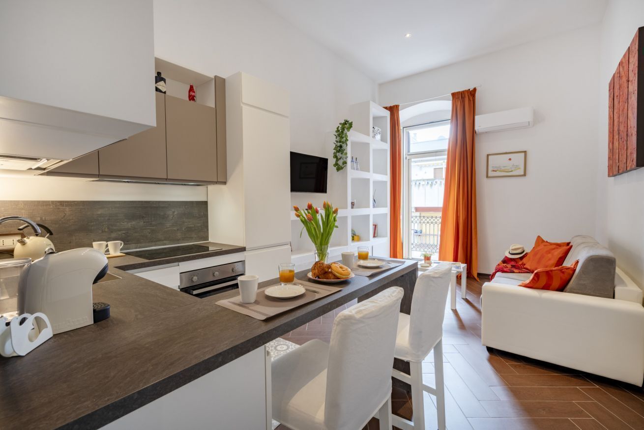 A modern kitchen with a central island, white furniture, and a dining area with orange curtains.