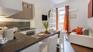 A modern kitchen with a central island, white furniture, and a dining area with orange curtains. - GuestHost - Lovely apartment for 4 people, located just 200 meters from the sea. The accommodation boasts every comfort e a palette of warm colors that create a welcoming and cheerful environment. Ideal location: just 5 minutes walk from Corso Vittorio E (Bari)