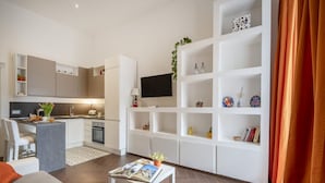 A modern living room with a sofa, a coffee table with magazines, a kitchen area with a TV, and a bookshelf with decorative items. - GuestHost - Lovely apartment for 4 people, located just 200 meters from the sea. The accommodation boasts every comfort e a palette of warm colors that create a welcoming and cheerful environment. Ideal location: just 5 minutes walk from Corso Vittorio E (Bari)