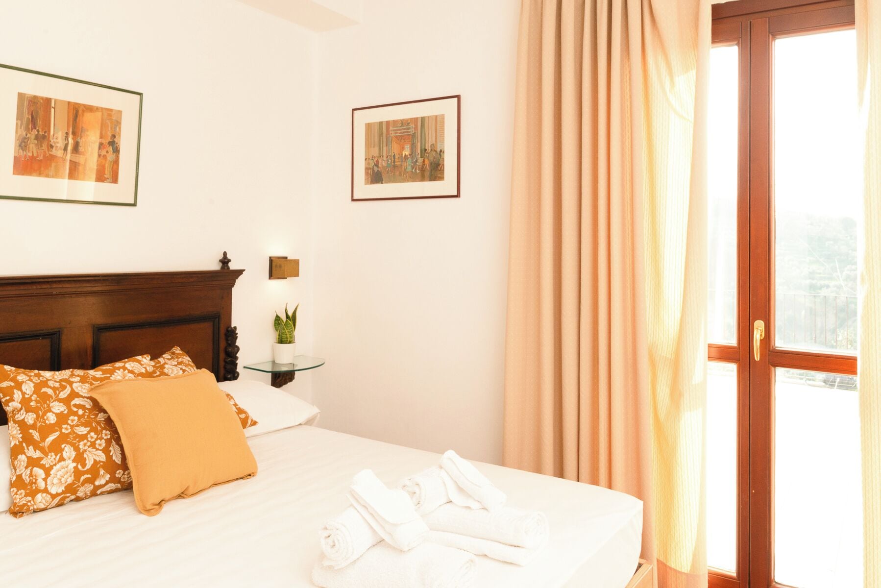 Une chambre d’hôtel avec un lit, deux œuvres d’art encadrées, une table de chevet avec une lampe et une fenêtre avec des rideaux.