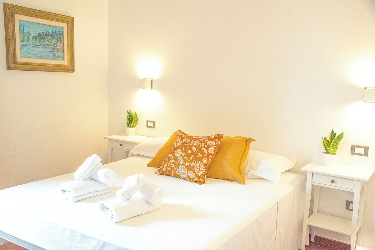 A hotel room with a bed, white linens, a floral pillow, two bedside tables with lamps, and a framed picture on the wall.