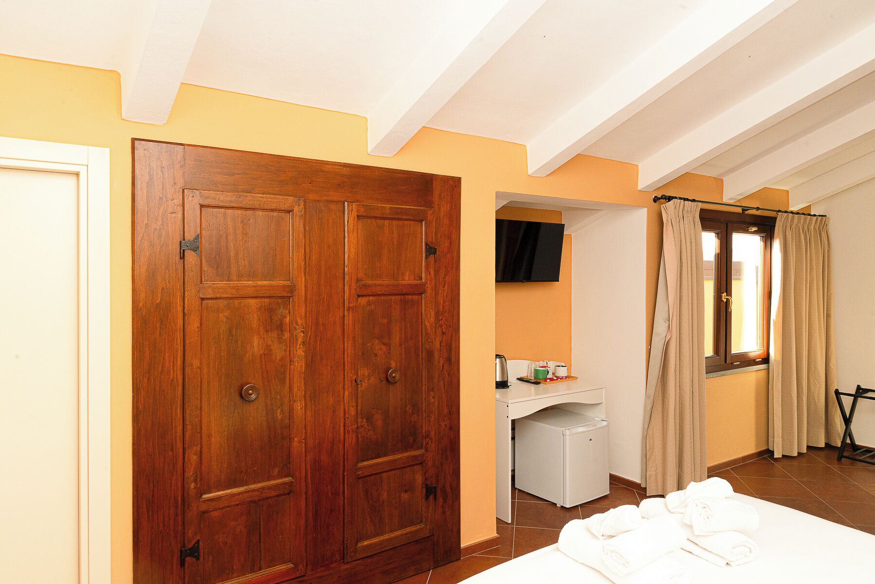 A room with a wooden door, a desk with a chair, a window with curtains, and a bed with towels.