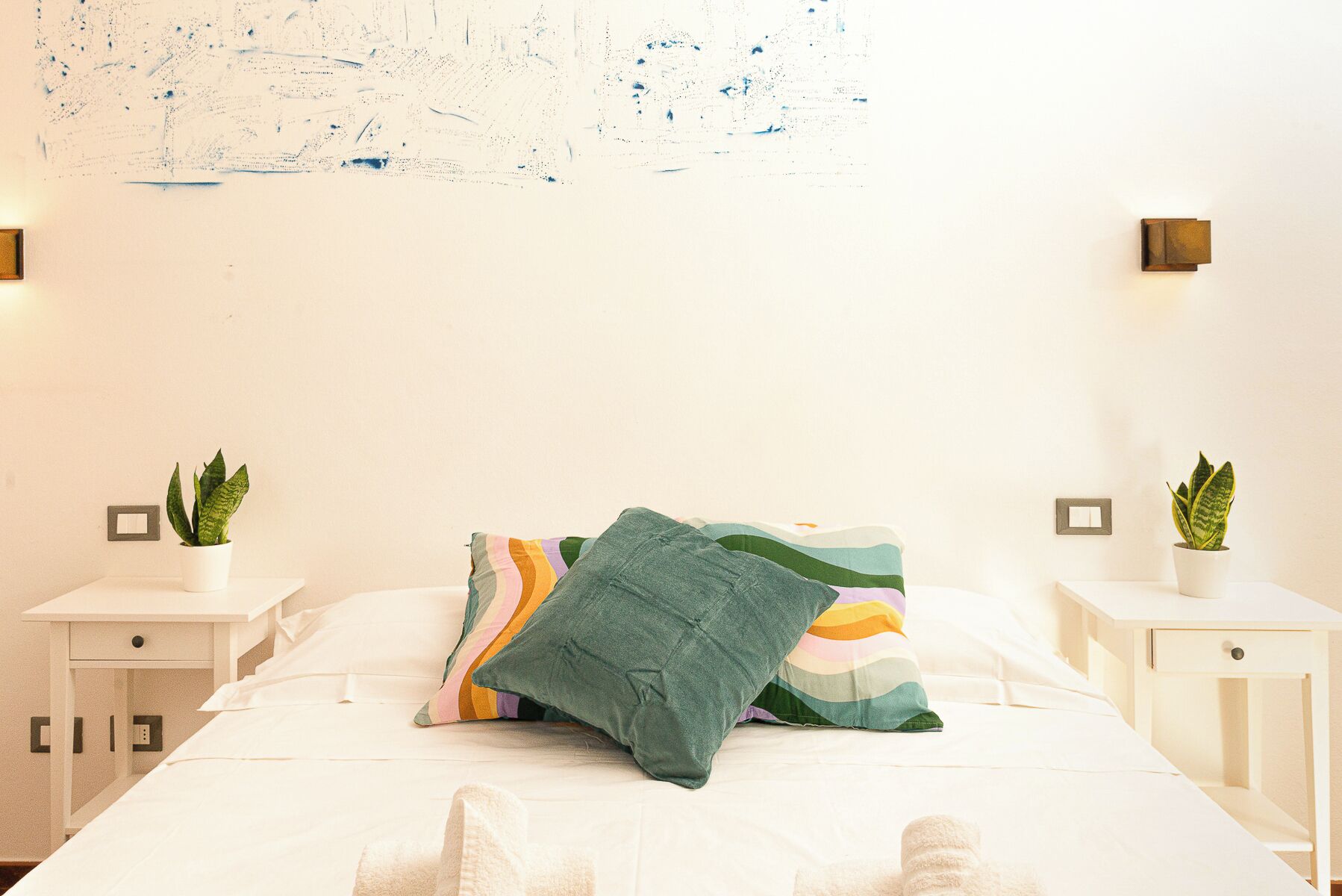 Una cama bien hecha con un cojín de colores, dos mesitas de noche con plantas en macetas y una pared con un cuadro abstracto con textura.