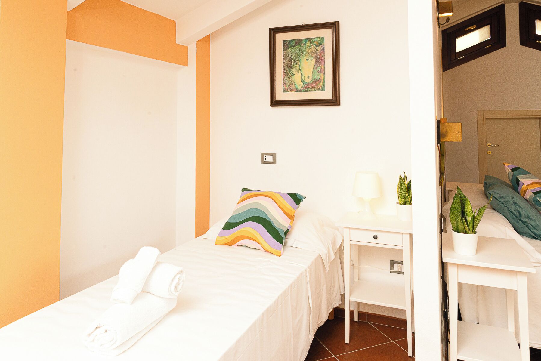A hotel room with a bed, a desk, a chair, a small table, a mirror, and a wall-mounted picture.