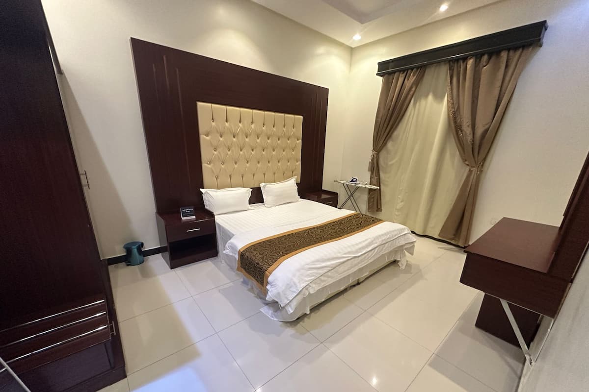 A hotel room with a large bed, a wooden headboard, a bedside table, a small table with a chair, and a window with curtains.