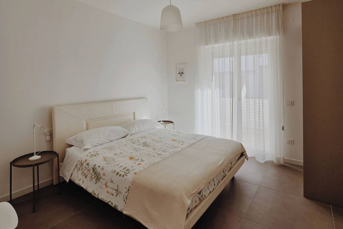 A bedroom with a large bed, a bedside table, a chair, and a window with curtains.