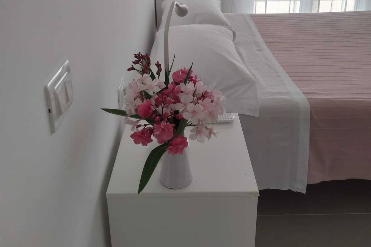 A hotel room with a bed, a white nightstand with a vase of flowers, a bedside table with a lamp, and a window with sheer curtains.