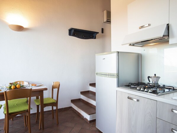 Una cocina compacta con un refrigerador blanco, una estufa y una cafetera. Hay una mesa de comedor con sillas y una pequeña escalera que lleva a un nivel superior.