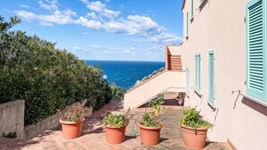 Un edificio costero con una escalera que lleva a un balcón con vista al mar, rodeado de macetas con plantas.