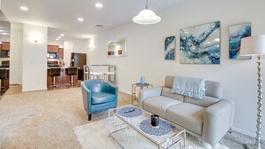 A modern living room with a beige sofa, a blue leather chair, and a glass coffee table. There is a kitchen in the background with a microwave and a refrigerator.