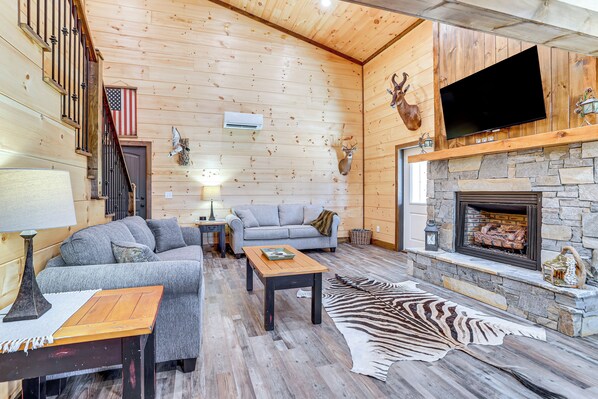 A log cabin interior with a stone fireplace, a mounted television, and a sofa. - Pet-Friendly Yard! Nature Escape in Forest City (Forest City)