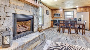 A stone fireplace with logs, a dining area with wooden chairs, a kitchen with a microwave and refrigerator, and a wooden ceiling. - Pet-Friendly Yard! Nature Escape in Forest City (Forest City)