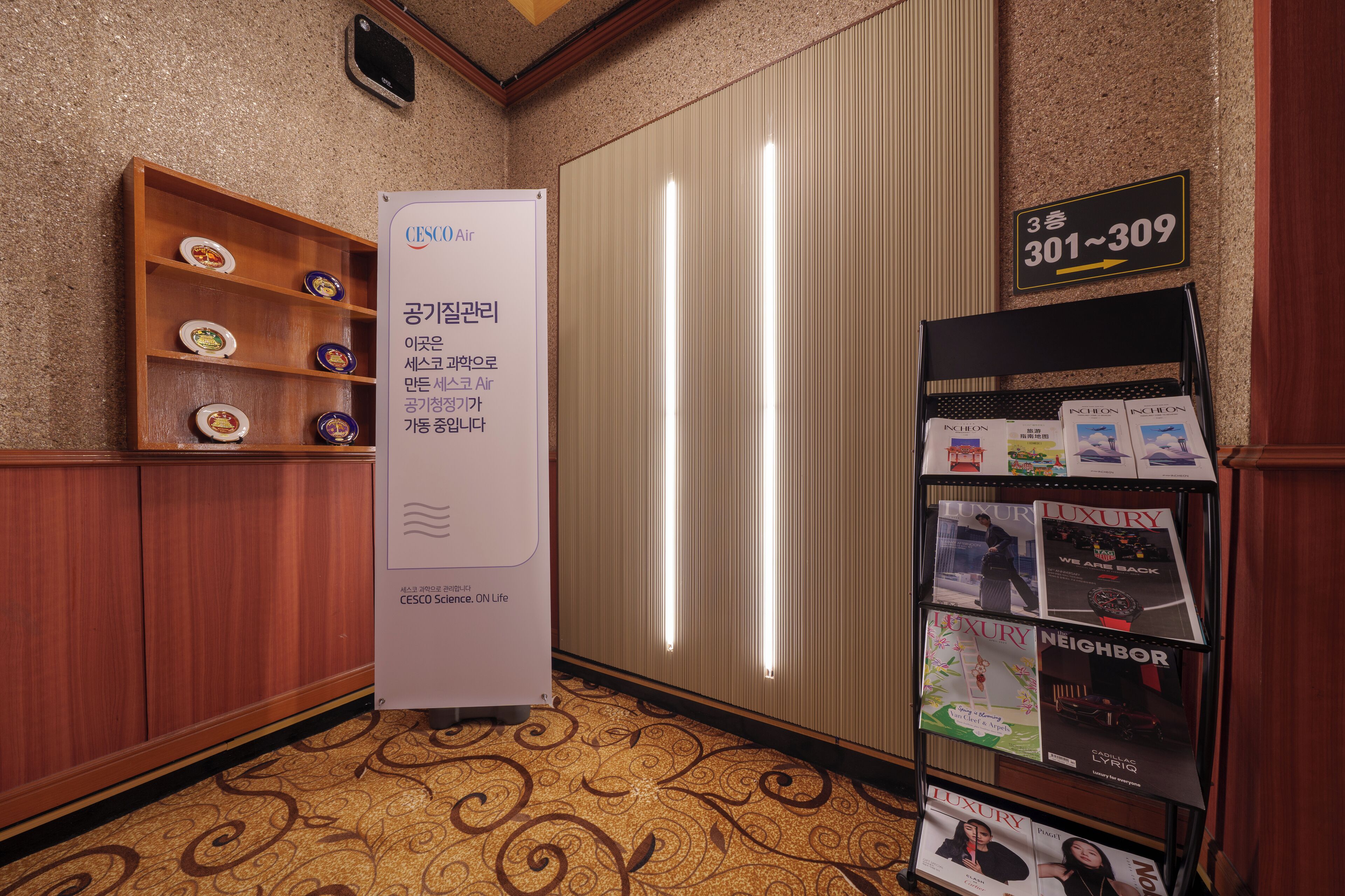 A hotel corridor with a wooden shelf displaying plates, a whiteboard with text, and a magazine rack with 'LUXURY' and 'NEIGHBOR' magazines.