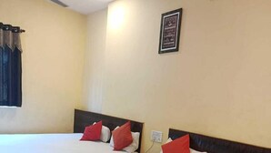 Une chambre d’hôtel avec deux lits simples, chacun doté d’une tête de lit en bois et de coussins rouges. On y trouve un ventilateur de plafond, une fenêtre avec des rideaux et un tableau encadré accroché au mur.