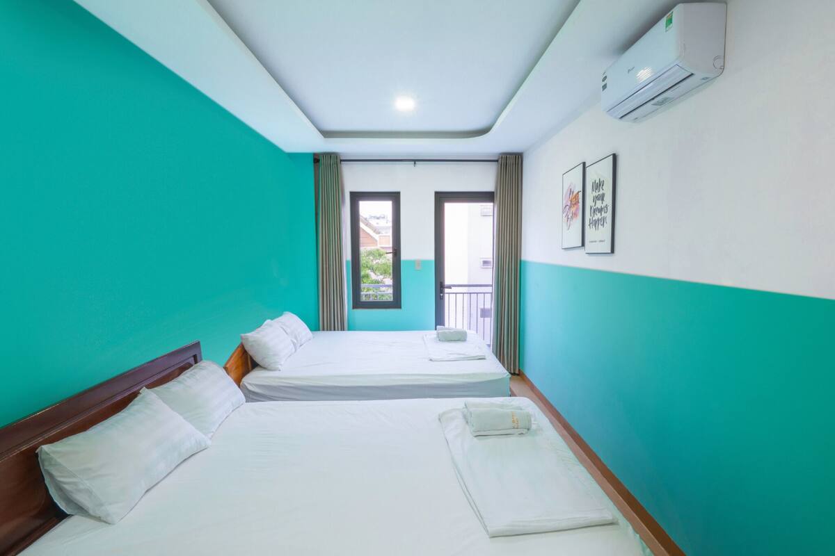 A hotel room with two beds, teal walls, a window with curtains, and an air conditioning unit.